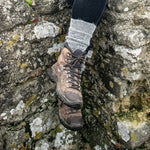A grey marl alpaca boot sock cuff turned down over a brown leather hiking boot, resting on a mossy stone wall.