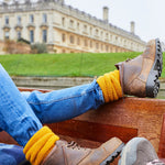 A person in jeans and hiking boots with mustard yellow alpaca boot socks turned over the boot cuffs, seated in front of a stately home.