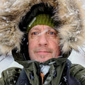A man taking a selfie in the snow wearing an olive green winter parka with a large brown faux-fur hood trim, a green knit beanie, and a black backpack, with a snowy train station platform and a blue train in the background.