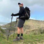A hiker in shorts and black alpaca crew socks standing with trekking poles on a grassy coastal clifftop path.