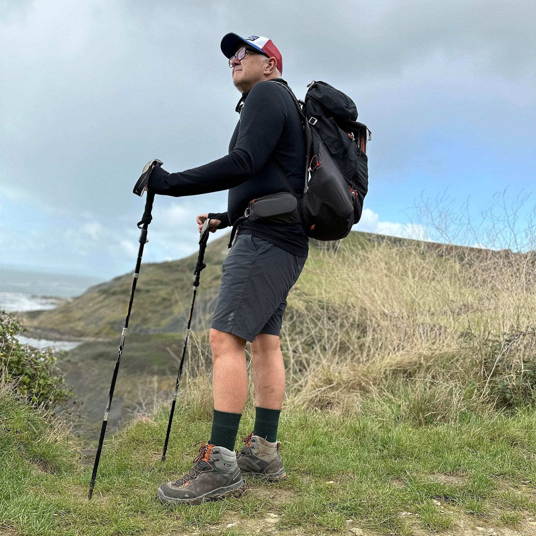 A hiker in shorts and black alpaca crew socks standing with trekking poles on a grassy coastal clifftop path.
