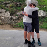 Two walkers in shorts and black alpaca crew socks embracing on a country lane beside a rocky bank.