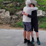 Two walkers in shorts and black alpaca crew socks embracing on a country lane beside a rocky bank.