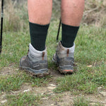 A walker's legs in dark green alpaca crew socks turned over grey leather hiking boots on rough grass.