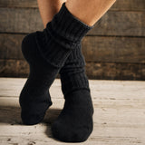 A close-up of a person's feet wearing black ribbed knit socks, standing on a light wood floor against a dark rustic wooden plank background.
