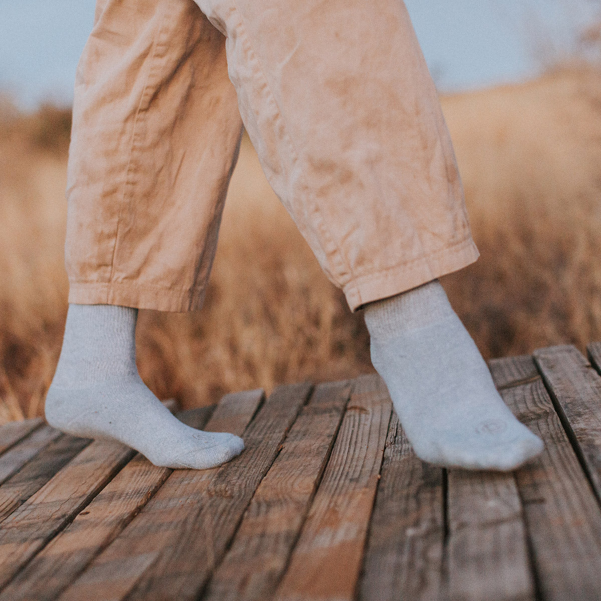 A close-up of a person's legs wearing light gray crew socks and tan pants, standing on a wooden boardwalk outdoors with dry grass in the background.