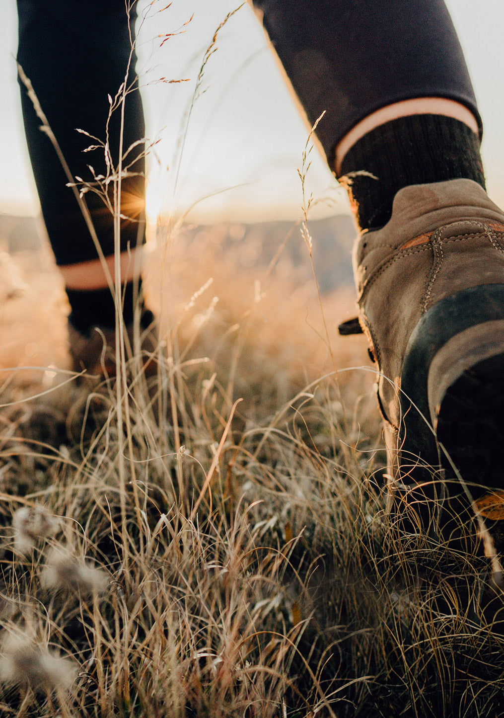 Close-up of a person hiking through dry grass at sunset, wearing dark leggings, black socks, and brown hiking boots.