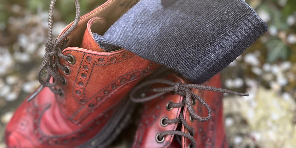 A close-up shot of a red leather lace-up boot with a dark gray knit sock tucked into the opening, set against a blurred outdoor background of green foliage and light-colored stones.