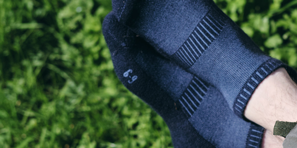 A close-up of a person's foot wearing a dark navy blue knit sock with ribbed stripes and a small white '6-9' marking, set against a blurred green grass background.