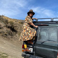 A woman wearing a brown wide-brim hat and a colorful floral midi dress stands on the back of a dark green off-road vehicle, looking back towards the camera in an outdoor desert landscape under a blue sky.