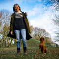 A woman stands outdoors on a grassy hill with bare trees and a blue sky, smiling while wearing a dark coat, patterned sweater, jeans, and boots. A brown dog sits nearby holding a tennis ball.