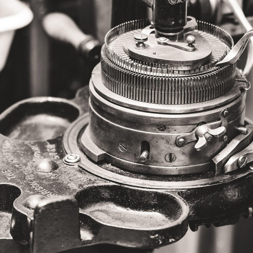 Close-up of a vintage mechanical typewriter or printing device with metal gears, levers, and a circular dial, shown in a dark monochrome photograph.
