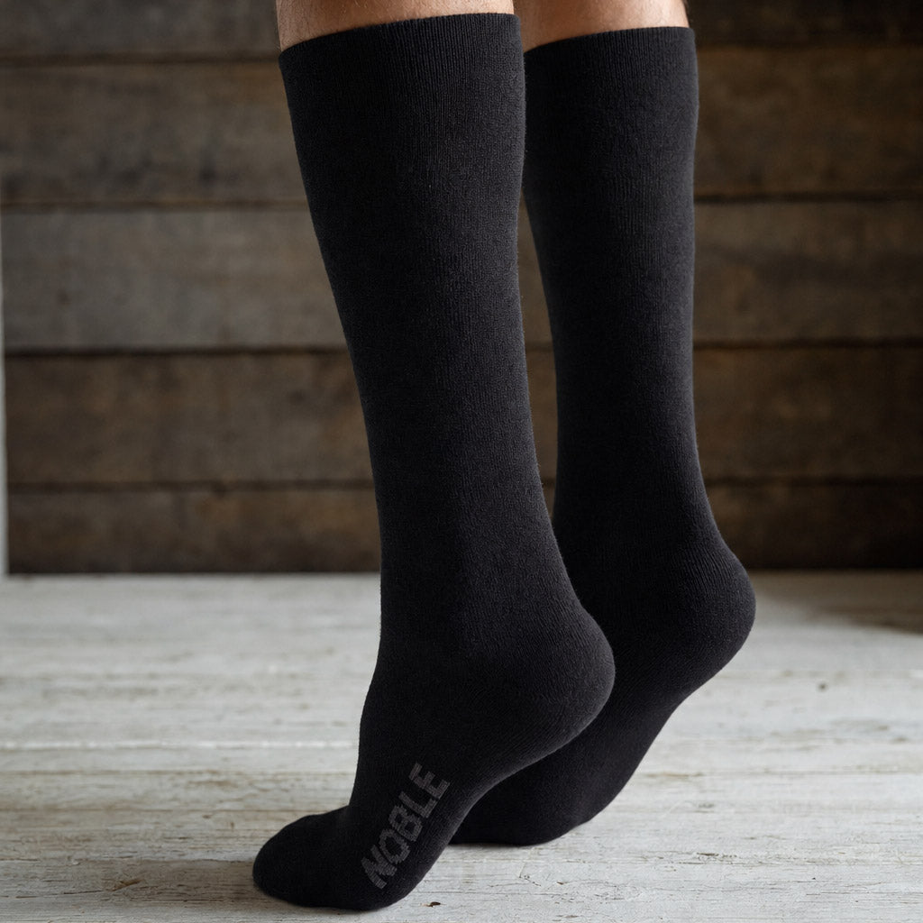 A pair of black crew socks worn on feet, with the brand name 'NOBLE' printed in gray on the sole of the front sock, set against a rustic wooden background.