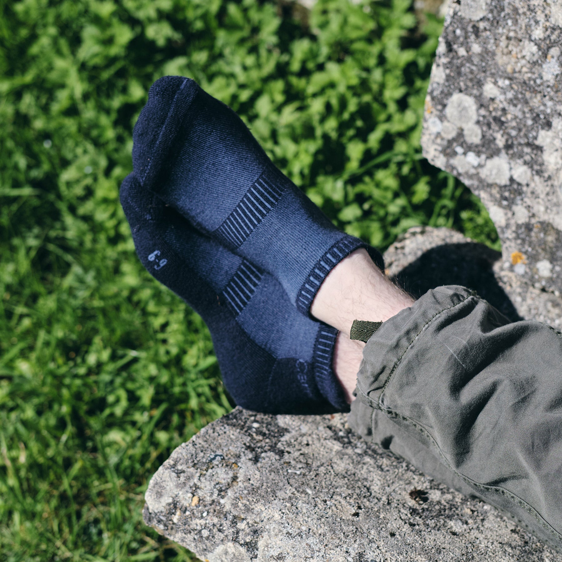 A navy merino trainer sock worn on a foot resting against a lichen-covered rock in green undergrowth.
