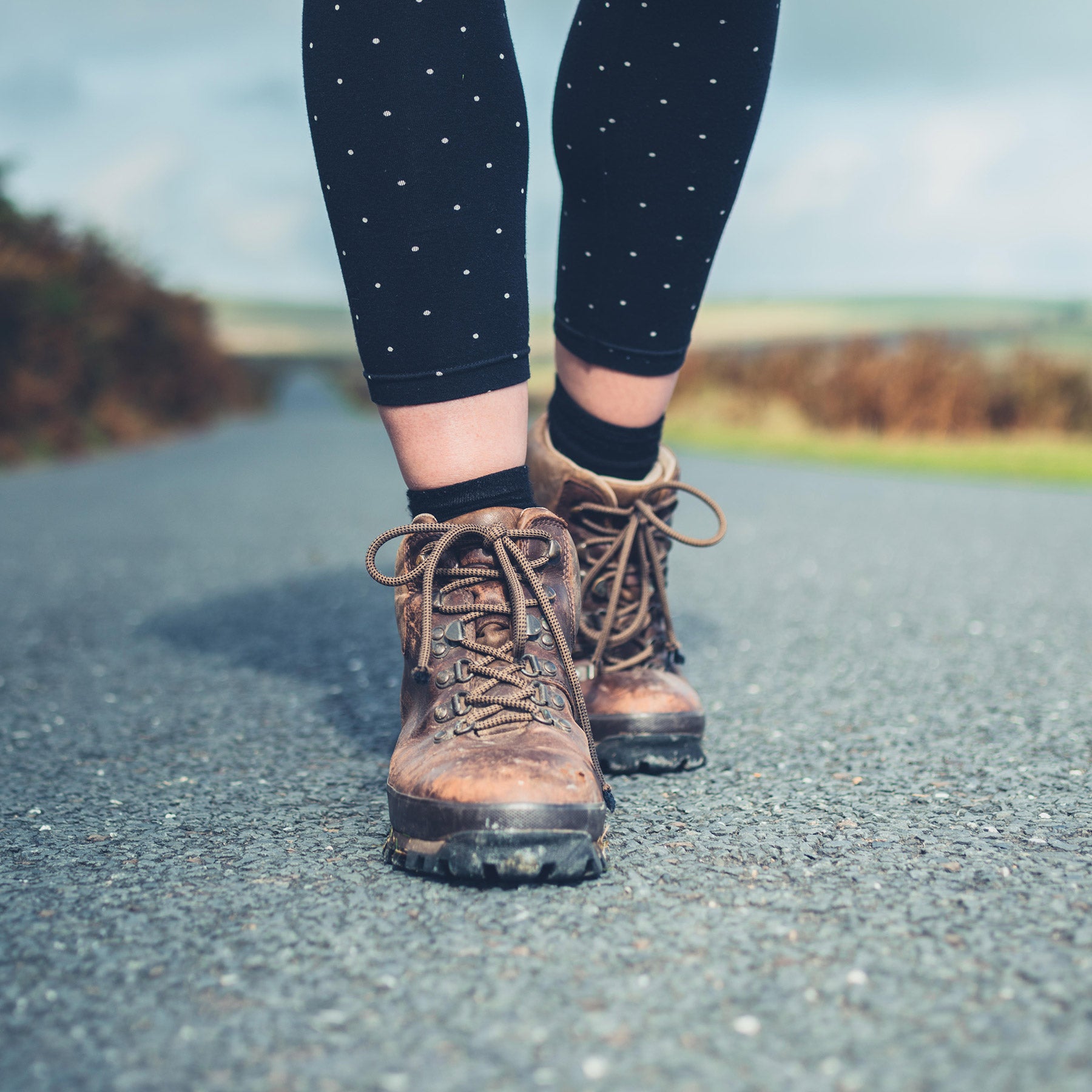A walker's legs in navy spotted leggings and muddy brown hiking boots on a tarmac road, wearing alpaca no-show socks hidden inside the boots.
