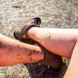 Mud-splashed bare legs in walking sandals resting on a rock, showing how alpaca no-show socks stay hidden.
