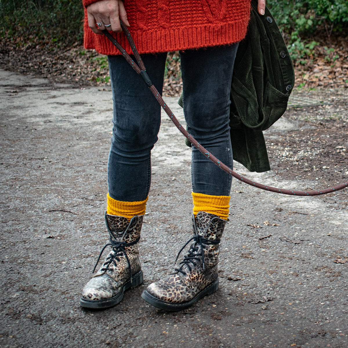 A person outdoors wearing leopard-print combat boots with yellow ribbed cuffs, dark skinny jeans, and a red knit sweater, holding a dark green jacket and a leather strap.