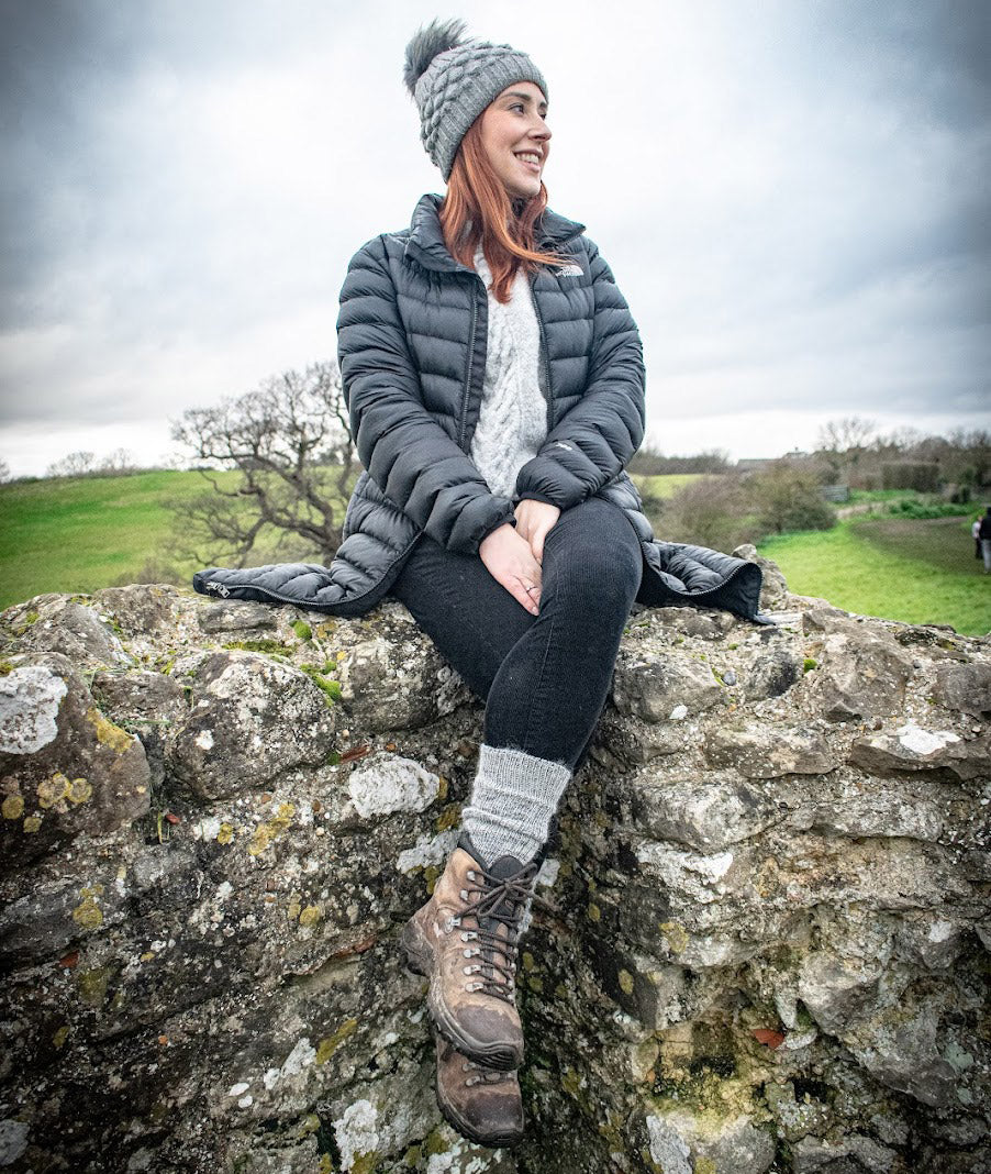 A woman wearing a dark gray puffer jacket, gray knit beanie with a pom-pom, black leggings, and brown hiking boots sits on a stone wall outdoors under an overcast sky.